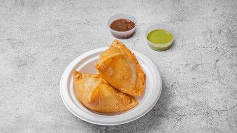 Two golden fried samosas on a white plate with bowls of red chutney and green sauce on a gray textured surface