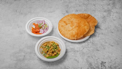 Indian meal spread with two fried breads on a plate, alongside small white bowls of vegetable curry and fresh onion and tomato garnish on gray surface